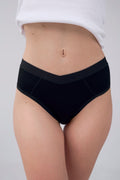 A close-up of a model's lower torso wearing black high-waisted briefs with a wide black waistband, paired with a white ribbed top against a light gray studio background.